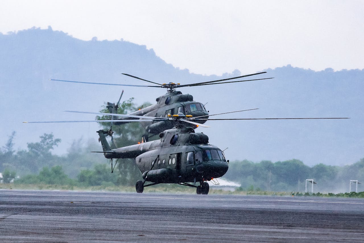 Two military helicopters on the runway during daytime, poised for takeoff in a rugged terrain.